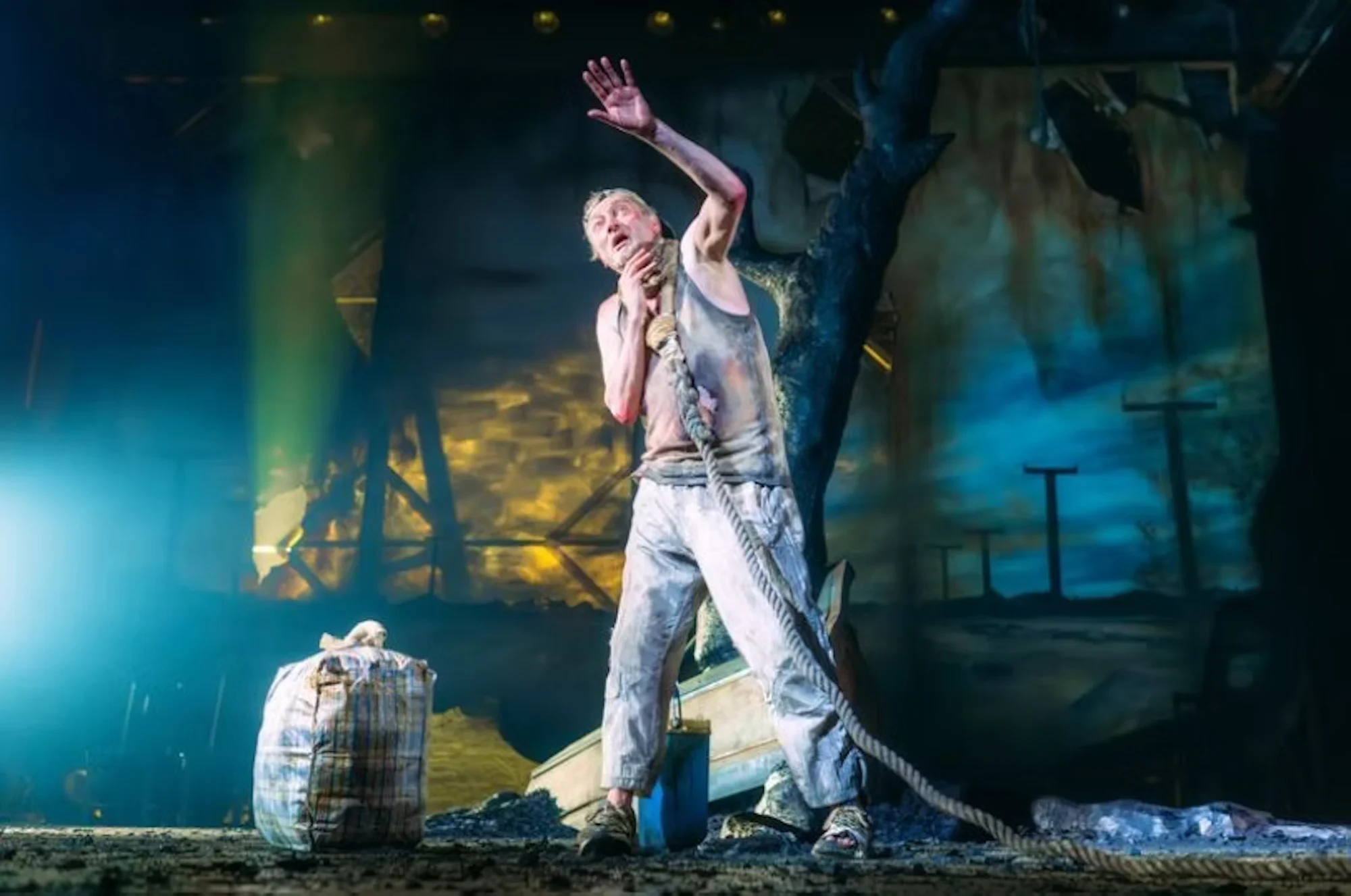 A man in tattered clothes clutches a rope on a dimly lit stage, exuding desperation. Beside him is a weathered bag, and a dramatic backdrop suggests a stormy setting.