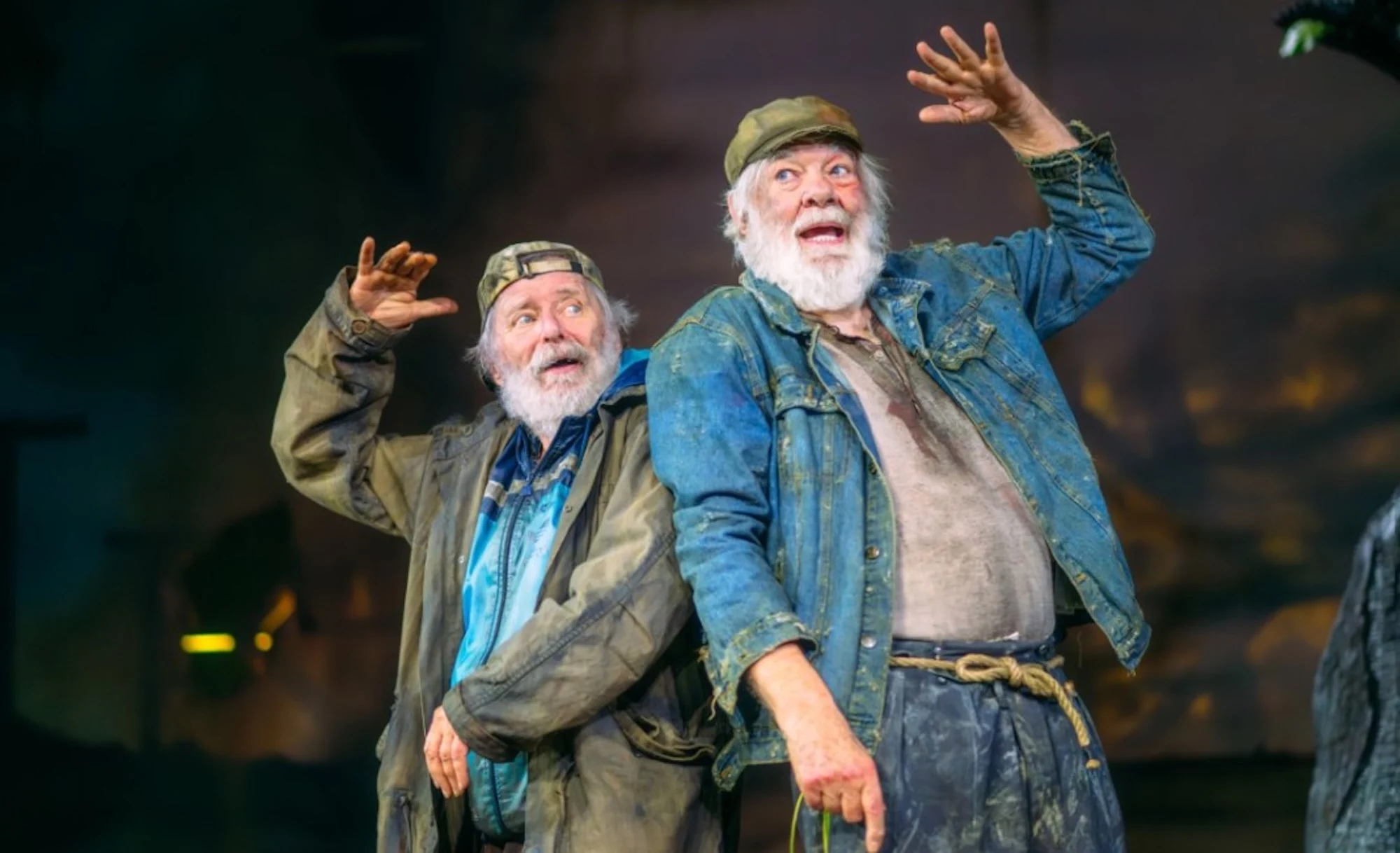 Two elderly men in worn, casual clothes, with joyful expressions, raise their arms playfully. The background is a softly lit, hazy setting.