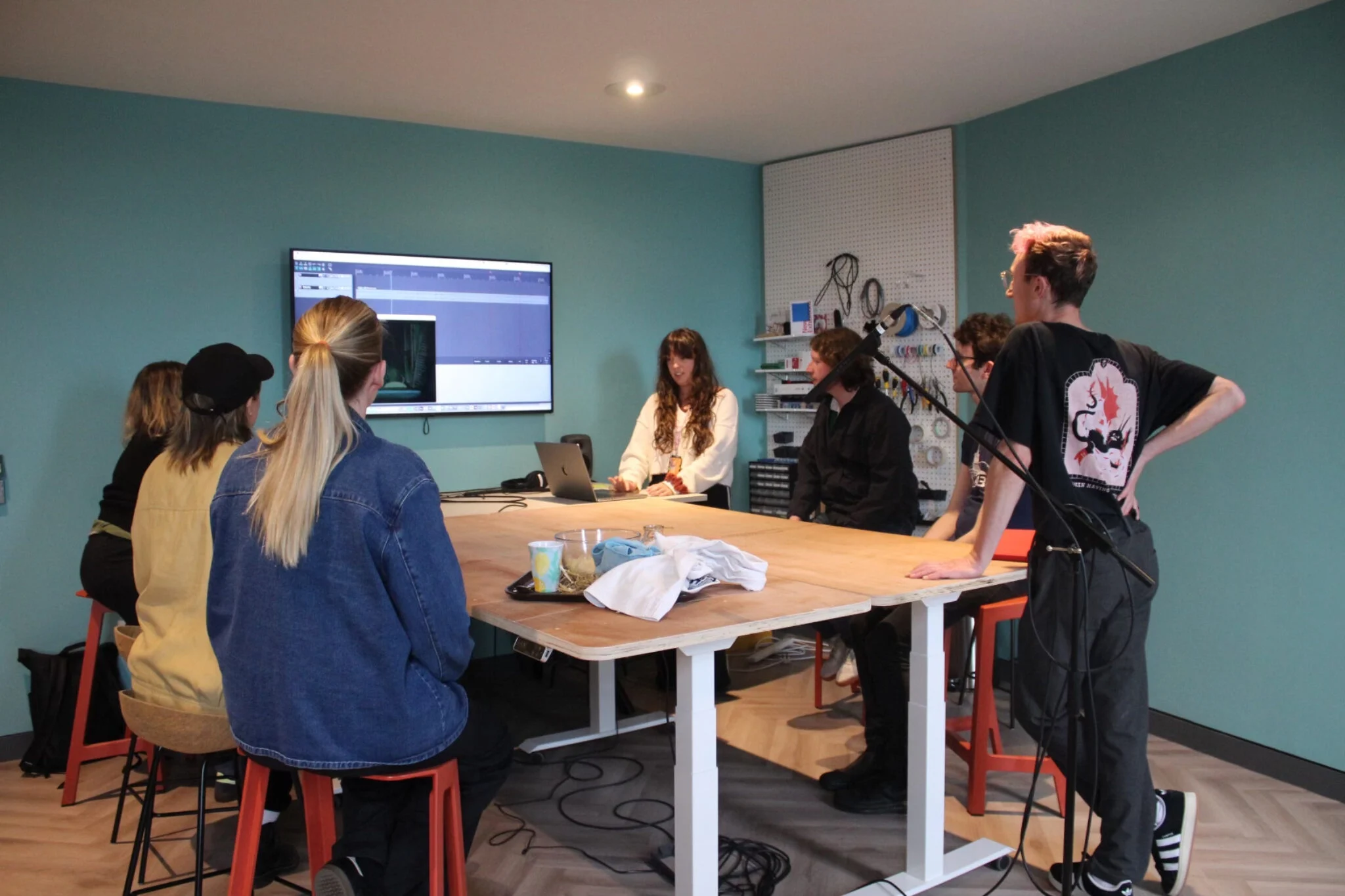 A group of people some sat and some standing around a table, talking